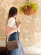 Bandolera color visón, fabricada en cuero con forro de tela. Tiene bolsillo interior y exterior. Entra billetera grande.