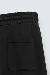 Pantalón jogger negro de corte relaxed fit, confeccionado en tejido de algodón. Presenta cintura elástica ajustable con cordón, pliegues frontales, bolsillos delanteros y un bolsillo trasero.