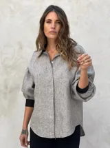 Camisa a rayas verticales grises y blancas, de lino, con cuello clásico, cierre frontal con botones, mangas abullonadas 3/4 con puños elásticos negros y corte holgado.