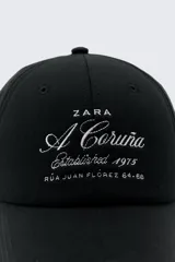Gorra de béisbol negra de Zara con bordado frontal en blanco que dice "A Coruña Established 1975 Rúa Juan Flórez 64-66".