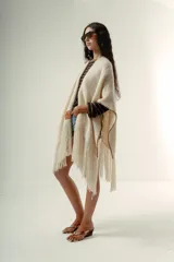 Ruana artesanal de lana merino color crudo, con tejido grueso y flecos largos en el borde. Presenta un ribete de costura en color marrón oscuro que bordea todo el contorno.