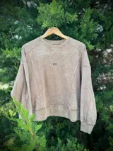 Buzo de algodón y poliéster color beige con efecto lavado (acid wash). Presenta un corte corto y holgado, con costuras decorativas verticales en el frente y un pequeño bordado de dos hojas en el pecho.