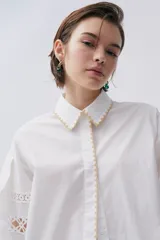 Camisa blanca de popelina con calce recto y levemente holgado. Mangas largas con galones florales y bordes festoneados en beige.