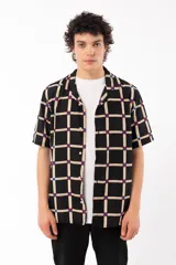 Camisa de manga corta con calce relajado y cuello tipo sastre, confeccionada en rayón. Presenta un diseño de cuadrícula beige sobre fondo negro con detalles en azul y rojo en las intersecciones.