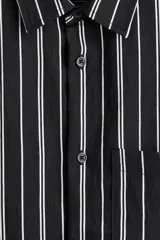 Camisa de manga corta con rayas verticales blancas sobre fondo negro, cuello inglés, tapeta con botones y bolsillo de parche en el pecho. Corte recto y holgado.