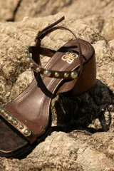 Sandalia de cuero color marrón con taco bloque medio y punta cuadrada. Presenta tachas esféricas doradas en las tiras y hebilla de ajuste en el tobillo.