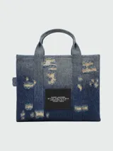 Bolso tote Marc Jacobs de tamaño mediano confeccionado en denim azul con efecto desgastado. Presenta el logo "THE TOTE BAG MARC JACOBS" estampado en blanco en el frente, doble asa corta superior y correa bandolera extraíble de tela negra.