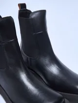 Bota Chelsea de cuero sintético color negro, con elástico lateral y tirador trasero.