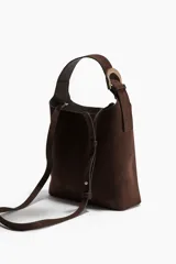 Bolso tipo hobo o bucket de textura cepillada (similar a gamuza o nobuk) color marrón oscuro. Posee un asa de hombro ajustable con una hebilla metálica dorada y una correa adicional ajustable para llevar cruzada.