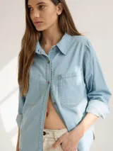 Camisa celeste de denim suave, corte relajado, con cuello clásico, cierre frontal con botones marrones, manga larga con puños abotonados, dos bolsillos de parche en el pecho y ruedo redondeado.