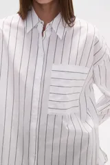 Camisa de algodón blanca de corte holgado, con cuello camisa y manga larga. Presenta un estampado de finas rayas verticales negras y un bolsillo frontal con rayas horizontales.