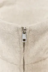 Cazadora corta color marrón claro, de cuello alto y manga larga. Tiene bolsillos laterales y cierre frontal con cremallera metálica.