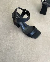 Sandalias negras de cuero sintético, con tira delgada sobre los dedos, pulsera al tobillo con hebilla metálica y taco cuadrado.