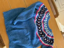 Sweater tejido a mano en lana mohair color crudo, con cuello redondo y mangas largas. Presenta un diseño con motivos geométricos en tonos marrón y verde en la parte superior del pecho.