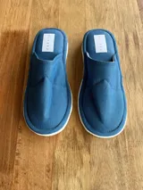 Pantuflas de tela acolchada color azul con suela blanca y etiqueta con el logo de la marca.