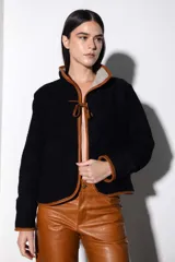 Chaqueta corta de paño a cuadros en tonos beige y negro, con cuello mao y ribete de cuero marrón en todo el contorno. Se ata con lazos de cuero en el centro y tiene bolsillos laterales.