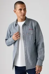 Camisa de manga larga a rayas verticales azules y blancas, con cuello americano, cierre frontal con botones a presión, dos bolsillos con solapa abotonada en el pecho y logo bordado en rojo sobre el bolsillo derecho.