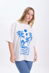 Remera blanca de corte amplio con estampado de palmeras, cabaña y aves en color azul.
