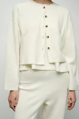 Chaqueta color crema de punto canalé con cuello redondo, manga larga y cierre frontal con botones marrones.