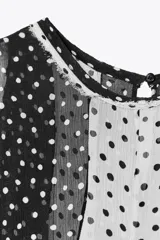 Top de cuello redondo y manga sisa con detalle de tejido plisado semitransparente y estampado de lunares blancos y negros. Bajo asimétrico y cierre en espalda con abertura y botón.