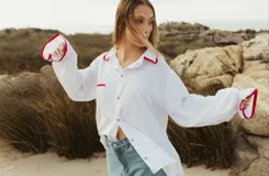 Camisa blanca de corte holgado con cuello camisero y mangas largas con puños elásticos. Presenta bolsillo de parche en el pecho y detalles de ribete rojo en cuello, puños y bolsillo.