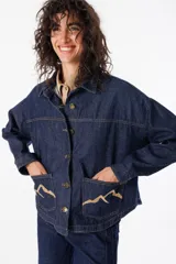 Camisa corta de jean azul con terminación redondeada en el ruedo. Presenta dos bolsillos plaqué bordados con motivo de montaña y detalle de pespunte a contratono.
