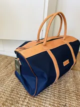 Bolso de lona azul con detalles en cuero marrón, doble asa de mano y correa bandolera ajustable. Incluye un neceser haciendo juego con cierre y logo de caballo bordado.