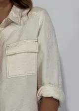 Chaqueta color gris con textura, de corte recto, cuello camisero, cierre frontal con botones, mangas 3/4 con puños arremangados, dos bolsillos de parche en el pecho y ruedo recto.