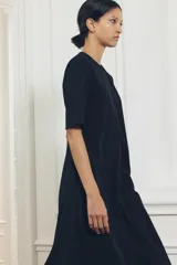 Vestido midi negro de cuello redondo y manga corta, con bolsillos laterales ocultos y costuras marcadas. Tiene corte evasé con abertura en la espalda y cierre trasero con cremallera oculta.