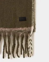 Bufanda tejida color crema con flecos en los extremos y etiqueta de cuero sintético negra.