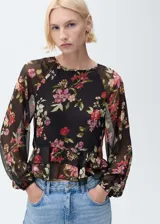 Blusa de mujer con estampado floral en tonos rojos, rosas y verdes sobre fondo negro. Tiene cuello redondo, mangas largas abullonadas, volantes en el bajo y cierre de lazo en el cuello.