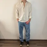 Camisa de manga larga color beige claro, de corte regular, con cuello clásico y cierre frontal con botones. Presenta un pequeño logo bordado en el pecho.