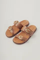 Sandalia plana de cuero color caramelo, estilo 'slide' o 'dos tiras', con hebillas ajustables de metal dorado en cada tira.