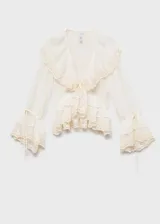 Blusa color crema con cuello en V, volados y detalles de encaje. Tiene mangas largas con puños elásticos y lazos.