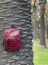 Mochila color bordó con textura tipo cuero de reptil en la parte superior y diseño capitoneado en la parte inferior. Tiene un bolsillo frontal con cierre y tiras regulables.