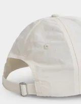 Gorra de gabardina color crudo con visera rígida y aplique ovalado de silicona color lila con texto.