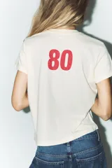 Camiseta color crudo de algodón con efecto lavado, cuello redondo, manga corta con hombro caído y estampado frontal con la leyenda "Texas USA Ranch Original est. 1975" en color rojo.