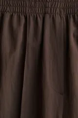 Pantalón marrón de corte holgado en sarga de mezcla de lyocell. Modelo de tiro alto con cintura fruncida, cierre decorativo y bolsillos laterales discretos. Piernas rectas con pliegues cosidos adelante y raya planchada atrás.