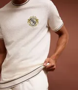 Remera de algodón y poliéster color crudo, con cuello redondo y manga corta. Presenta textura acanalada horizontal, estampa localizada con escudo y ribetes en contraste en cuello y mangas.