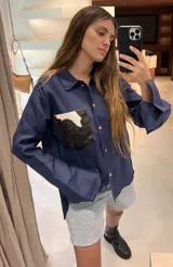 Camisa color naranja de lino o algodón con cuello clásico, cierre frontal con botones y mangas largas con puños abotonados. Presenta un bolsillo de cuero marrón en el pecho.