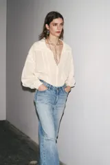 Camisa color crudo confeccionada en hilatura con mezcla de ramio, de corte holgado, con cuello solapa y escote pico. Tiene mangas largas con puños y detalle de tejido con pliegues.
