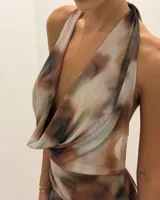 Vestido largo de tela estampada con efecto tie-dye o marmolado en tonos marrones, beige y negro. Presenta un escote halter con cuello drapeado tipo cascada y un corte ajustado al cuerpo.