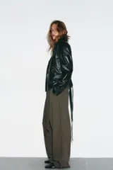 Chaqueta negra de efecto piel con cuello solapa, manga larga y trabillas en los hombros. Presenta bolsillos delanteros y cierre frontal con botones, ajustada a la cintura con un cinturón del mismo tejido.