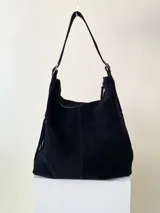 Bolso tipo hobo color tostado, hecho de cuero con flecos largos en el frente y dos bolsillos con cierre. Tiene una manija regulable con hebillas metálicas.