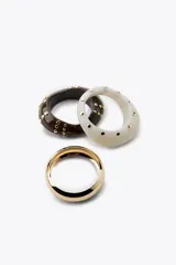 Set de tres pulseras rígidas: una dorada de metal y dos de resina, una color crema y otra marrón, ambas con tachas doradas.