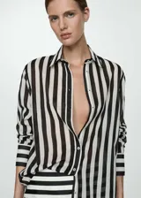 Camisa de manga larga con puños abotonados, cuello camisero y cierre frontal con botones. Diseño a rayas verticales blancas y negras sobre tejido semitransparente.