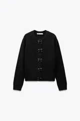 Chaqueta de punto negra con cuello redondo y manga larga. Presenta detalle de lazos con abalorios en el frente y cierre con botones a presión.