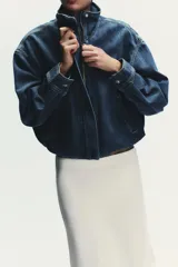 Chaqueta corta de denim azul con cuello alto, manga larga con hombros caídos, bolsillos laterales, bajo con elástico y cierre frontal con cremallera y botones ocultos.