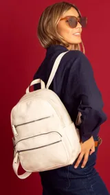 Mochila color crema de símil cuero con tres bolsillos frontales con cierre. Tiene asa de mano rígida y correas regulables en galón a tono con hebillas metálicas.