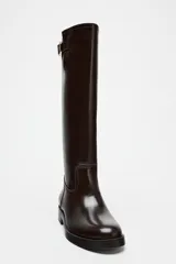 Bota alta de cuero color marrón oscuro, con caña alta, punta redonda y hebilla decorativa en la parte superior.